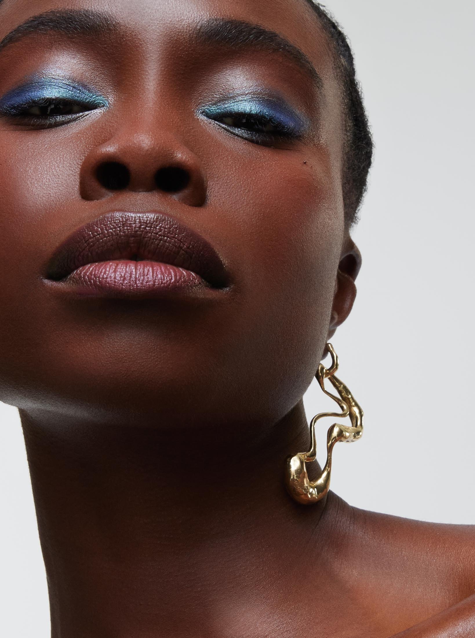 Close-up studio shot of a dark-skinned woman with blue eye shadow wearing a gold earring, set against a neutral grey background.